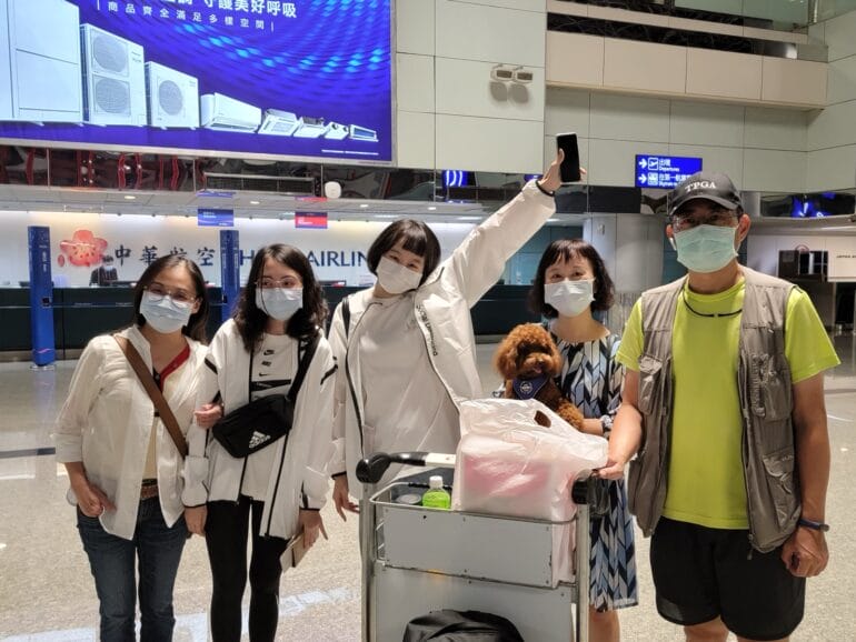 五名戴口罩的人在機場與小狗及行李推車合照，中間的人舉起手機