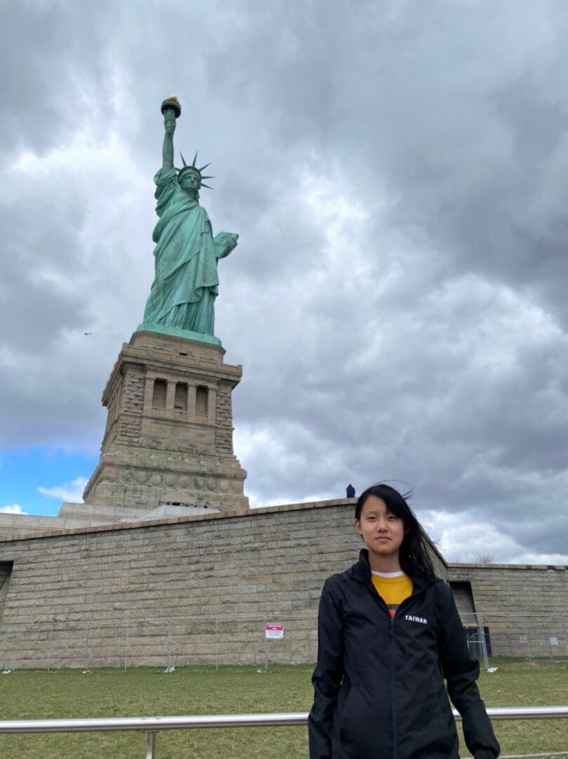 一名穿深色外套的學生站在自由女神像前,背景是多雲天空